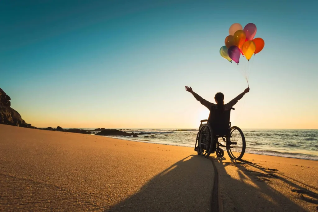 A woman in a wheelchair holding balloons with both arms in the air sits on a beach watching the sunrise.